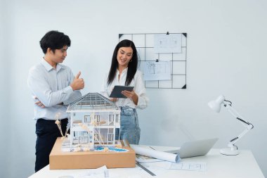 Güzel, genç, beyaz bir proje yöneticisi Asyalı mühendise, mühendislik masasında ev modelini ölçerken tablet kullanarak bina detaylarını anlattı. Tertemiz.