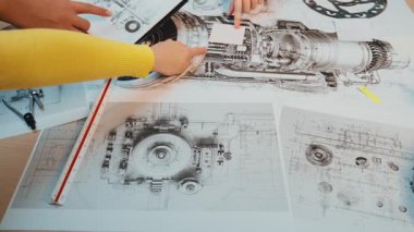 Bir el, detaylı mühendislik şemaları ve çalışma alanına yerleştirilmiş teknik çizimlerle meşgul olur. Araçlar ve bileşenler görünür, işbirlikçi bir ortam yaratıyor. SACTR