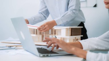 Ev modelinin uzunluğunu ölçmek için cetvel kullanan profesyonel erkek mimarın yakın plan görüntüsü. Bu arada genç iş arkadaşı ev modeliyle toplantı masasındaki verileri analiz etmek için dizüstü bilgisayar kullanıyor. Tertemiz..