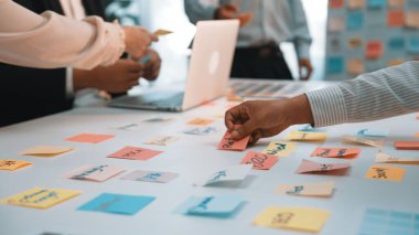 Dinamik bir ofis sahnesinde, bir dizüstü bilgisayarın etrafında beyin fırtınası yapmak ve fikir üretmek için renkli yapışkan notlar kullanarak, ortak bir toplantıda yer alan bir ekip görülüyor. SACTR