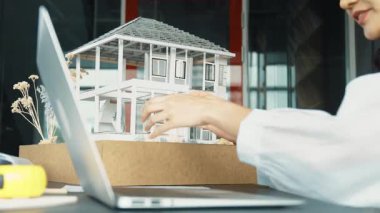 Genç ve güzel mimar mühendisin çatı malzemelerini kontrol etmesi ve ofisteki masaya yerleştirilmiş proje planı ve ev modeliyle dizüstü bilgisayarı kullanması. Yaratıcı iş tasarımı konsepti. Yönlendirici.