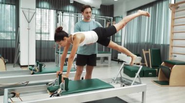 Bir pilates eğitmeni, müşteriye bir ıslah makinesinin tahta varyasyonunda rehberlik eder. Sağlıklı bir kadın, dengeyi geliştirmek için antrenör yardımı ile yenilikçi üzerinde yan kalas değişimi yapıyor. Alışkanlık.