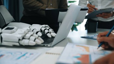 Dizüstü bilgisayarın yanında robot bir el gösteren modern bir çalışma alanı, proje tartışması, veri analizi ve yaratıcılığı teşvik eden bir iş ekibiyle birlikte. Noogenesis