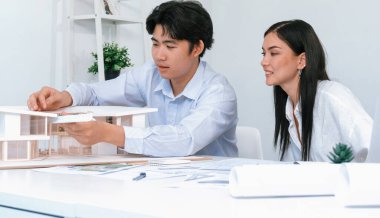 Profesyonel Asyalı erkek mimar ev modelinin uzunluğunu ölçmek için cetvel kullanırken, beyaz tenli genç meslektaşı ev modeliyle toplantı masasındaki verileri analiz etmek için dizüstü bilgisayar kullanıyor. Tertemiz..