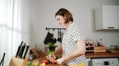 Enerjik anne, mutfak malzemeleri ile çevrili sebzeleri doğrarken modern mutfakta kahvaltı için salata yapıyor. Akıllı kadın temizlikçi yemek pişirir ve yemek hazırlar. Sağlıklı bir yaşam tarzı. Pedagoji.
