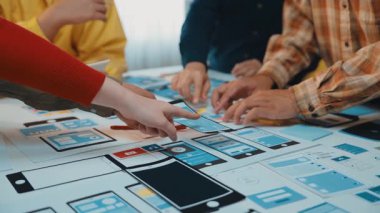 Çeşitli bireylerden oluşan bir grup, modern ofis ortamında arayüz tasarım stratejilerine odaklanarak renkli kağıt modellemeleri üzerinde işbirlikçi beyin fırtınası yapıyor. SACTR