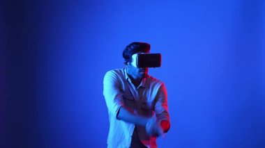 Meta-World 'de atış oyunu oynarken VR kulaklık takan profesyonel bir oyuncu. Erkek model, sanal gerçeklik gözlüğü kullanarak mavi arka planda dururken silah hareketi yapıyor. Aygıt.