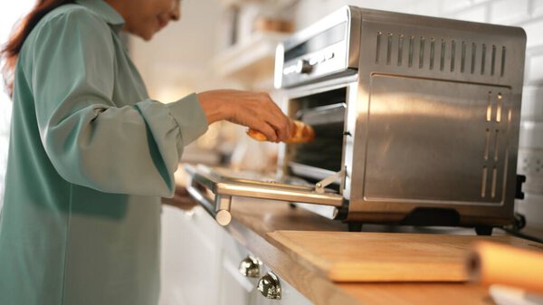 Закройте счастливую пожилую женщину, открывающую духовку и кладущую хлеб в духовку или пекущую хлеб. Счастливая бабушка готовится к завтраку, гуляя на домашней кухне и готовя еду на завтрак. Мирмидон.