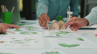 Çevresi yeşil elementler ve bilgilendirme çizelgeleri ile çevrili sürdürülebilir iş stratejileri üzerinde çalışan çeşitli bir grubu resmeden canlı bir ofis sahnesi. SACTR