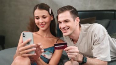 Genç çift, kredi kartıyla ödemek için internet üzerinden ödeme uygulaması ve dijital cüzdan kullanarak yatak odasında oturuyor. Mobil internet üzerinden alışveriş ve modern alımlar. Düzenle
