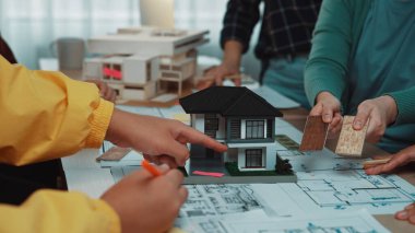 Bir grup mimar ve müşteri, mimari planlar ve malzeme örnekleriyle çevrili, takım çalışması ve yaratıcı keşiflerle çevrili bir ev modeli tasarlıyorlar. SACTR