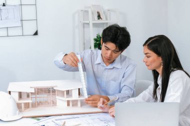 Profesyonel Asyalı erkek mimar ev modelinin uzunluğunu ölçmek için cetvel kullanırken, beyaz tenli genç meslektaşı ev modeliyle toplantı masasındaki verileri analiz etmek için dizüstü bilgisayar kullanıyor. Tertemiz..