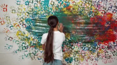 Resim dersinde sıradan beyaz gömlek giyerken, renkli bir elle boyalı duvarın arkasını gören güzel bir kız. Dekorasyon odası için öğrenci mührü. Yaratıcı eğitim konsepti. Öğretim