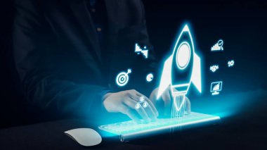 Bir işadamı, parlak bir klavyenin üzerinde, yenilik, dijital teknoloji ve modern bir çalışma alanında başarı hırsını temsil eden holografik bir roket sembolü ile meşgul olur. Dörtgen