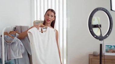 Genç sosyal medyum kadın moda videosu yapıyor. Blogcu kameraya selfie çubuğu ve ışık halkası kullanarak gülümserken, aynı zamanda ikna edici çevrimiçi kıyafetlerin izleyicilere veya takipçilere vlog satmasını sağlıyor. Düzenle