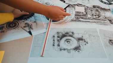 Ahşap bir masa üzerindeki mühendislik planları ve mekanik tasarımların detaylı bir görüntüsü, karmaşık çizimleri ve makine parçalarının teknik çizimlerini inceleyen elleri gösteriyor. SACTR