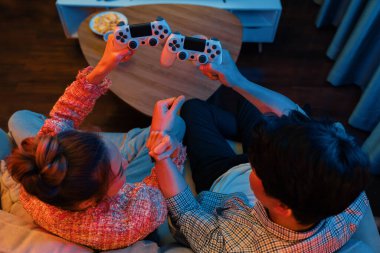 Çarpışan joystickleri olan, el ele tutuşarak kazanan, modern evdeki neon ışıklı oturma odasının arka tarafında video oyunu oynayan genç çift oyuncusunun en iyi görünümü. İnfobahn.