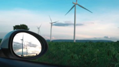 İlerleyen gelecek enerji altyapısı rüzgar türbini kavramı, rüzgar türbininin yeşil ve yenilenebilir enerjisiyle şarj edilen elektrik aracının yan aynasına yansıyor..