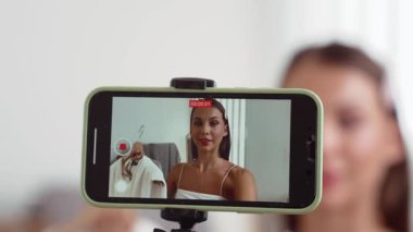 Genç sosyal medyum kadın moda videosu yapıyor. Blogcu kameraya selfie çubuğu ve ışık halkası kullanarak gülümserken, aynı zamanda ikna edici çevrimiçi kıyafetlerin izleyicilere veya takipçilere vlog satmasını sağlıyor. Düzenle