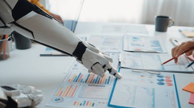 Gelecekçi bir robot kolu, bir ofis masasında insan eliyle iş raporları üzerinde işbirliği yaparak teknoloji ve insan çabasının veri analizlerindeki sinerjisini gösteriyor. Noogenesis