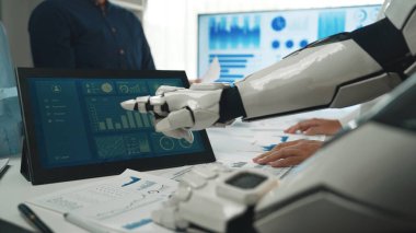 Bir robot modern ofis ortamında bir insan takımıyla işbirliği yapar, dokunmatik ekrandaki verileri analiz eder, teknolojilerin iş verimliliğindeki rolünü gösterir. Noogenesis