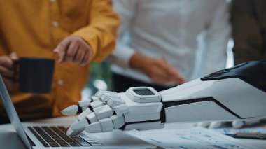 Bu resim, insanlar ve teknoloji arasındaki işbirliğini modern bir çalışma alanında sergiliyor, bir dizüstü bilgisayar ve takım üyeleriyle etkileşen robotik bir kol. Noogenesis