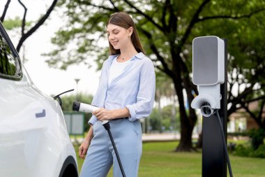 Açık yeşil şehir parkı manzaralı EV şarj istasyonundan genç kadın EV elektrikli araçları şarj ediyor. Çevre dostu şehir ulaşımı ve çevre dostu EV araba seyahati. Yükselt