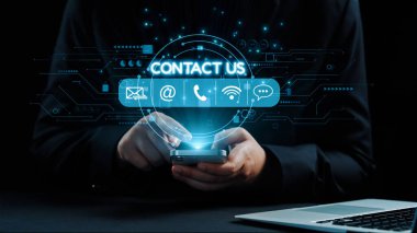 Dijital dünyadaki işletmeler ve bireyler için modern iletişim metotlarını sembolize eden, aydınlatılmış temas simgeleriyle akıllı bir telefonu tutan ellerin yakın çekimi. Dörtgen