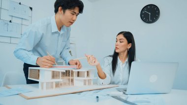 Profesyonel Asyalı erkek mimar ev modelinin uzunluğunu ölçmek için cetvel kullanırken, beyaz tenli genç meslektaşı ev modeliyle toplantı masasındaki verileri analiz etmek için dizüstü bilgisayar kullanıyor. Tertemiz..