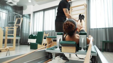 Eğitmen, müşteriye pilates reformatöründe rehberlik ediyor. Vücut geliştirme egzersizinde, vücut hareketlerine, esnekliğine ve beden kuvvetine odaklanmak için kayışlar kullanıyor. Asyalı kız pilates makinesinin önünde yatıyor. Alışkanlık.