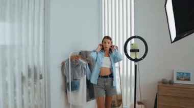 Genç sosyal medyum kadın moda videosu yapıyor. Blogcu kameraya selfie çubuğu ve ışık halkası kullanarak gülümserken, aynı zamanda ikna edici çevrimiçi kıyafetlerin izleyicilere veya takipçilere vlog satmasını sağlıyor. Düzenle