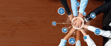 Ahşap bir masa üzerinde bir araya gelen çeşitli eller, dijital teknoloji ve ağ ikonlarıyla geliştirilmiş takım çalışmasını ve iş birliğini sembolize eder. Dostluk