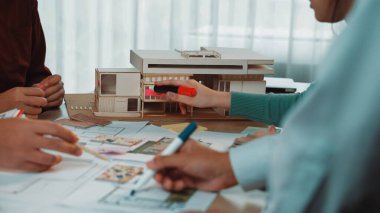 Bir grup mimar, eskizleri ve bir modeli inceleyerek modern bir çalışma alanında iş birliği yapıyor. Renkli kalemler ve tasarım kağıtları yaratıcı beyin fırtınası sürecini gösteriyor. SACTR