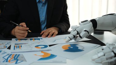 Bir işadamı, teknoloji ve insan zekasının modern ofis ortamında birleştirilmesini gösteren robotik bir elin yardımıyla mali raporları değerlendirir. Noogenesis