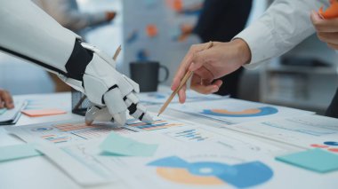 Modern bir ofis sahnesi, yenilikçi iş çözümlerini keşfetmek için veri analizleri, çizelgeler ve grafikler kullanan bir insan ve robot arasındaki işbirliğini gösteriyor. Noogenesis