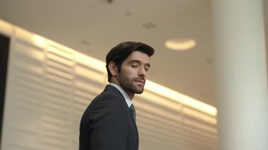 Profesyonel bir iş adamı kolları bağlıyken arkasını döner. Kafkasyalı mutlu yatırımcı, arka planı bulanık bir şekilde koridorda kollarını kavuştururken kameraya özgüvenle bakıyor. Portre. Sevinçli..