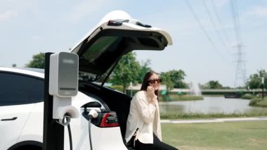 Yavaş çekimde genç bir kadın alışveriş merkezinin otoparkındaki şarj istasyonundan EV aküsünü şarj ederken akıllı phopne 'da konuşuyor. Modern kadın elektrikli araçla alışverişe gider. Yararlı