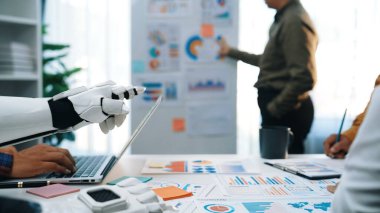 Modern bir ofis sahnesinde robot bir asistanın bir iş toplantısında insan takımıyla işbirliği yaptığı bir sahne. Takım üyeleri grafikler ve grafikler üzerinde sunulan verileri analiz eder. Noogenesis