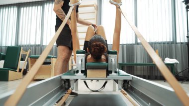 Eğitmen, müşteriye pilates reformatöründe rehberlik ediyor. Vücut geliştirme egzersizinde, vücut hareketlerine, esnekliğine ve beden kuvvetine odaklanmak için kayışlar kullanıyor. Asyalı kız pilates makinesinin önünde yatıyor. Alışkanlık.