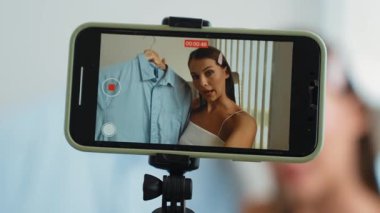 Genç sosyal medyum kadın moda videosu yapıyor. Blogcu kameraya selfie çubuğu ve ışık halkası kullanarak gülümserken, aynı zamanda ikna edici çevrimiçi kıyafetlerin izleyicilere veya takipçilere vlog satmasını sağlıyor. Düzenle