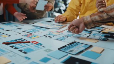 Çeşitli bir ekip, modern bir çalışma alanındaki yapışkan notlar ve dijital cihazlarla dolu bir masadaki kullanıcı arayüzü kablolarını inceleyerek, işbirlikçi bir tasarım oturumuna girişir. SACTR