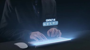 Pürüzsüz bir klavyeye yazı yazan ellerin yakın plan görüntüsü, karanlık bir arkaplan karşısında, e-posta ve telefon gibi ışıldayan dijital arayüz kontak seçenekleri sunuyor. Dörtgen