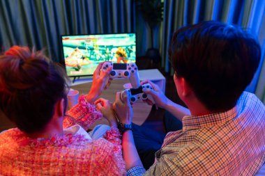 Kontrollü joystickli birkaç oyuncu arka tarafta televizyon ekranında dövüş oyunu oynuyor, modern rahat evinde neon ışıklı renkli oturma odasında koltukta oturuyor. İnfobahn.