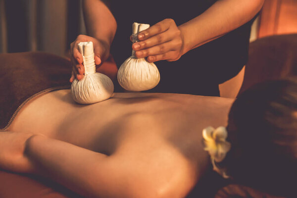 Hot herbal ball spa massage body treatment, masseur gently compresses herb bag on couple customer body. Serenity of aromatherapy recreation in warm lighting of candles at spa salon. Quiescent