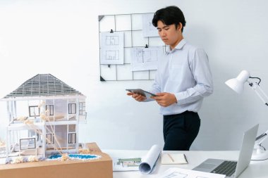 Profesyonel yakışıklı Asyalı proje yöneticisi ev modelini kontrol etmek için tablet kullanırken ev modelinin yapısını dikkatlice inceleyip arkasında planlar asılıydı. Yaratıcı tasarım. Tertemiz..