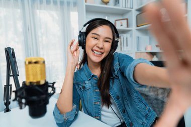 Sunucu kanalı genç ve güzel Asyalı kız akıllı telefonuyla selfie çekiyor. Modern stüdyo ofis konseptinde kulaklık takıyor. Sahne arkası yayıncısı sosyal medyada canlı yayın yapıyor. Taktik.