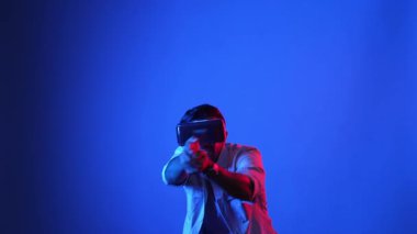 Meta-World 'de atış oyunu oynarken VR kulaklık takan profesyonel bir oyuncu. Erkek model, sanal gerçeklik gözlüğü kullanarak mavi arka planda dururken silah hareketi yapıyor. Aygıt.