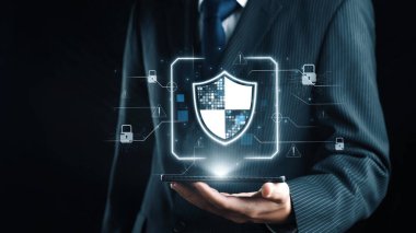 Siber güvenliği simgeleyen dijital kalkan grafiğini gösteren bir akıllı telefonu elinde tutan bir işadamı, çevrimiçi güvenlik ve veri korumanın önemini vurguluyor. Dörtgen