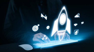 Modern bir çalışma alanında yenilik, büyüme ve teknolojiyi temsil eden holografik başlangıç simgeleriyle çevrili bir klavye ile iş yapan bir profesyonel. Dörtgen