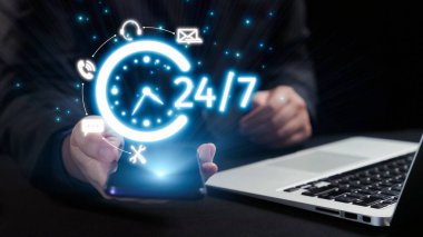 Bir dizüstü bilgisayarın yanında 24 saatlik servisi sembolize eden, profesyonel bir ortamda modern bağlantıyı ve ulaşılabilirliği gösteren akıllı telefonu gösteren canlı bir görüntü. Dörtgen